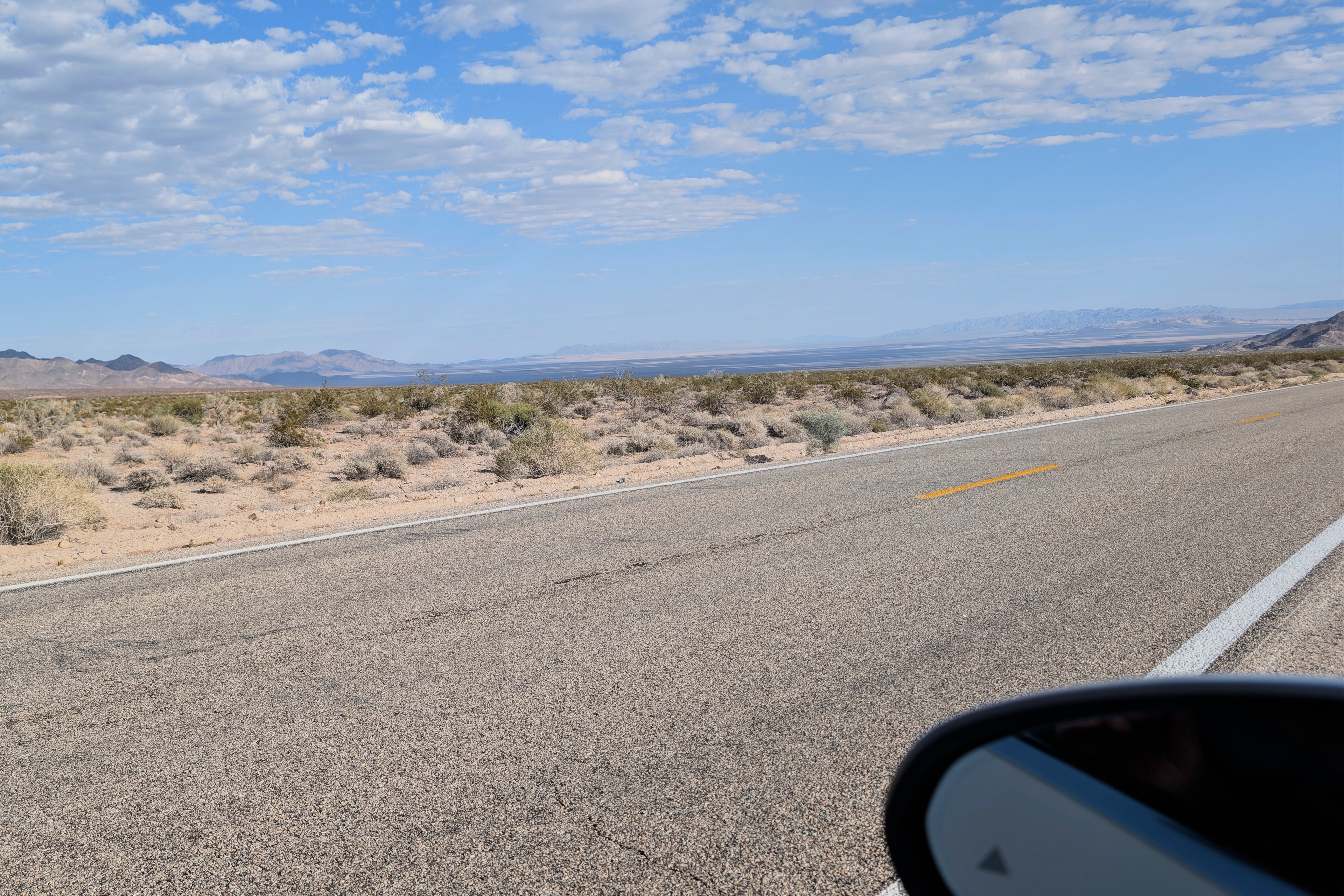 Two-lane road through the Mojave seen from the driver&rsquo;s seat, side mirror in the corner, distant mountains under scattered clouds