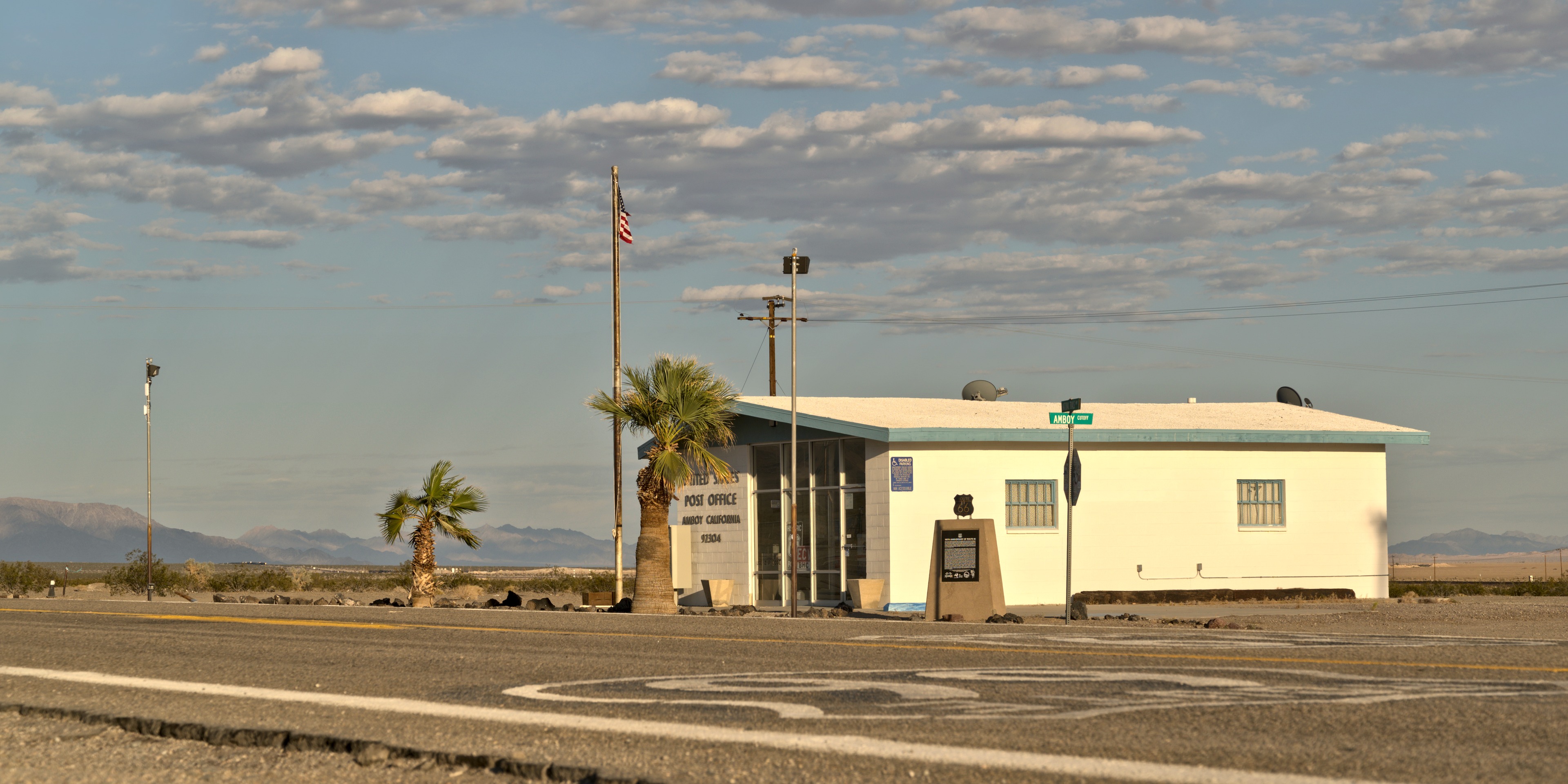 Amboy post office in afternoon light: low white building with blue trim, palm tree, US flag, Route 66 painted on the asphalt