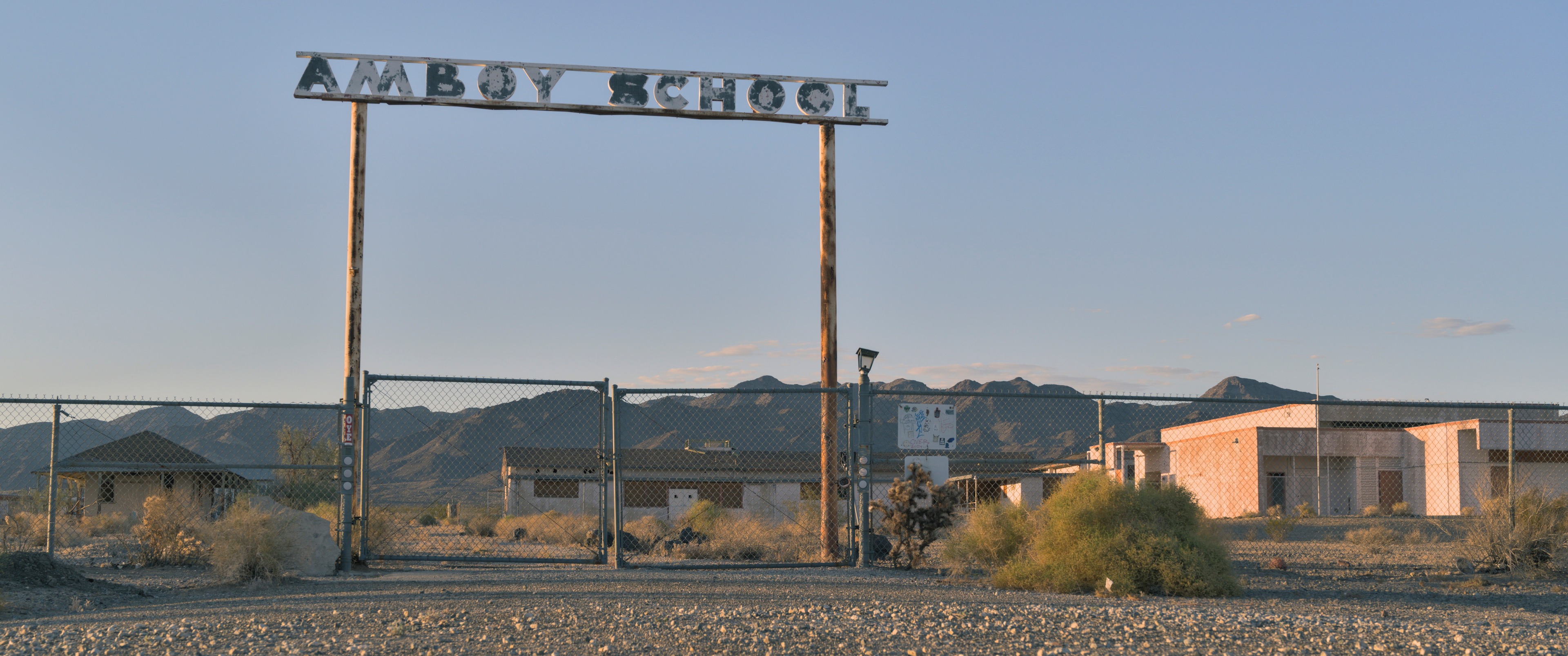 Abandoned Amboy School behind a chain-link gate, weathered school sign overhead, mountains beyond