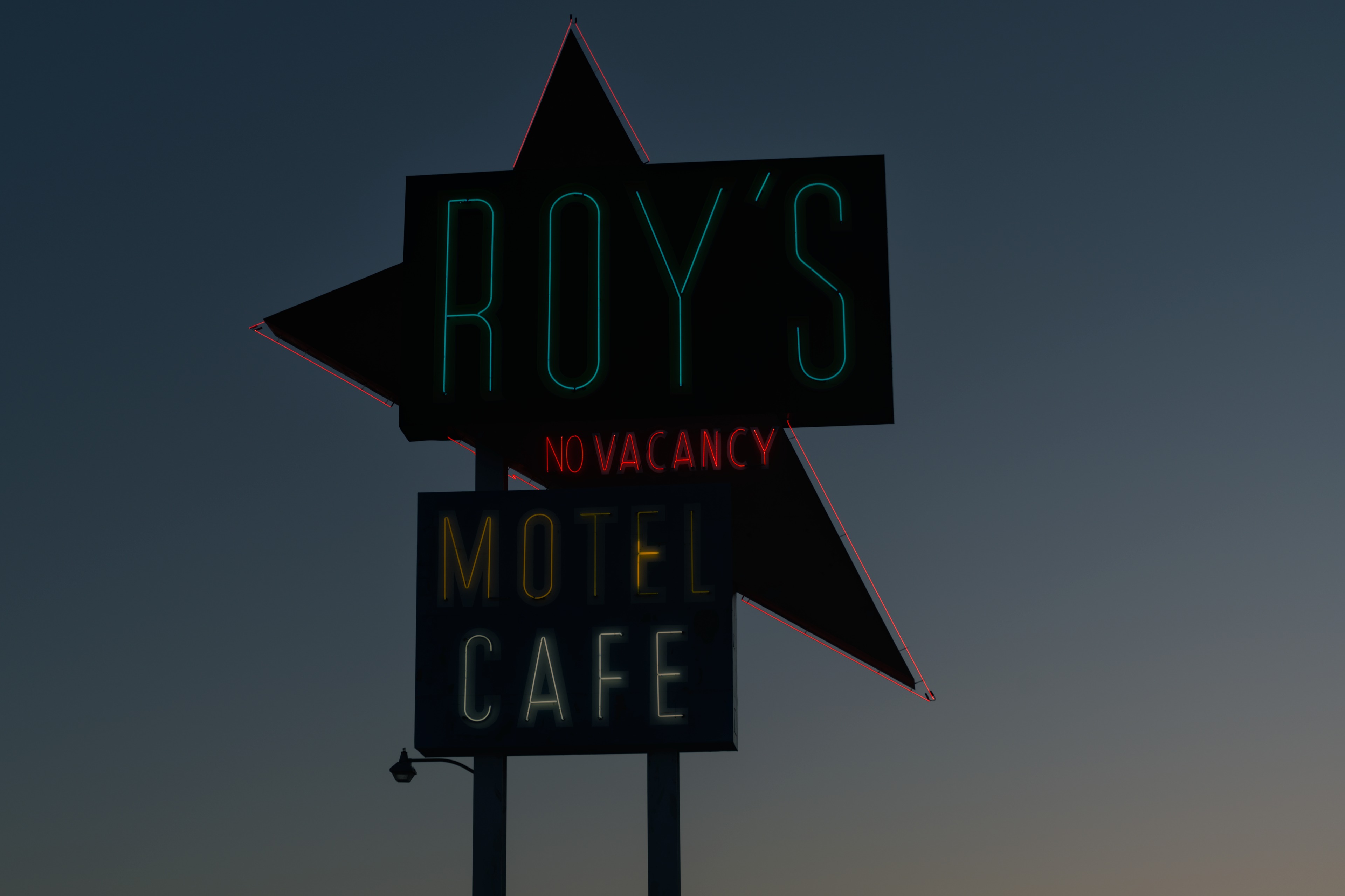 Roy&rsquo;s Motel &amp; Cafe neon sign at twilight: &lsquo;No Vacancy&rsquo; glowing red, &lsquo;Motel Cafe&rsquo; lit in yellow and white, deep blue sky fading to amber at the horizon