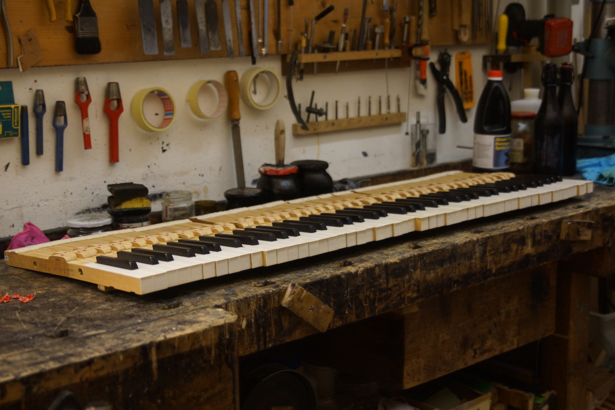A piano keyboard under restoration on a scarred wooden workbench, hand tools, punches and tape rolls hanging on the wall behind it