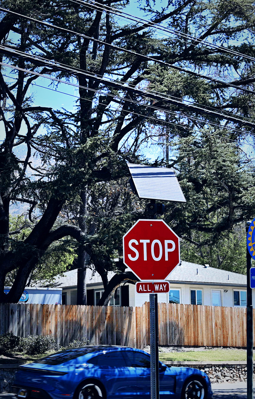 A Flock license-plate-reader camera mounted above an all-way STOP sign at a residential intersection, a car passing through below